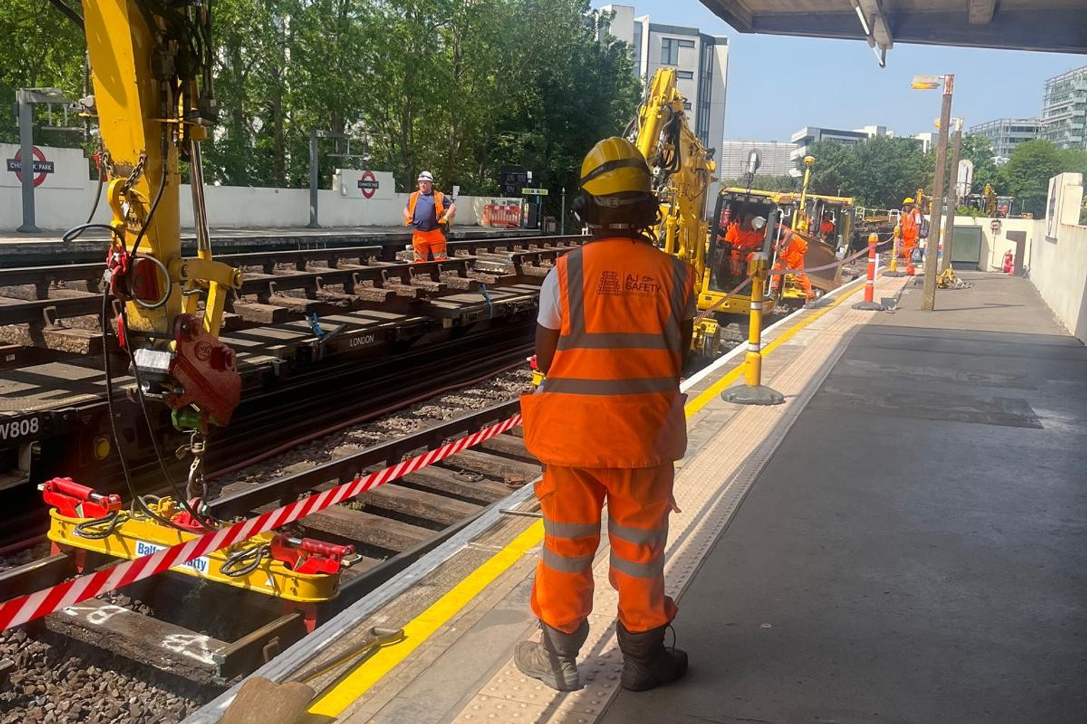 AJ trackside worker beside rail plant and a sleeper-handling attachment at an Underground platform