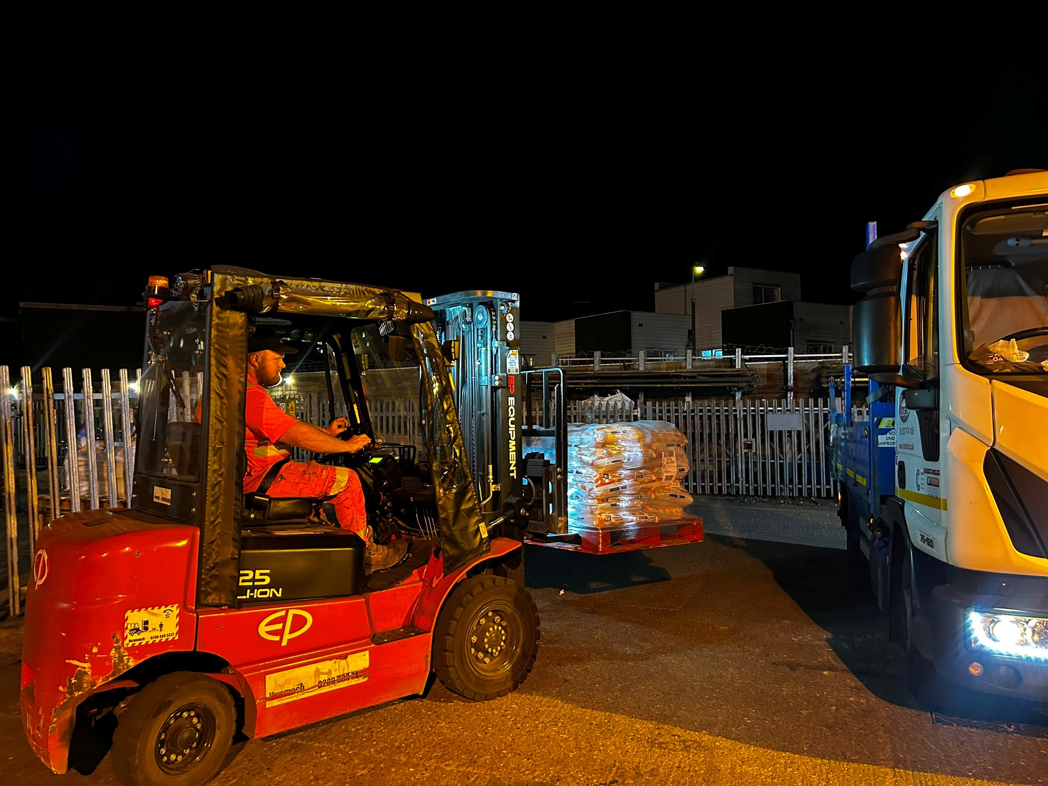 Forklift loading palletised materials beside a vehicle during a night shift