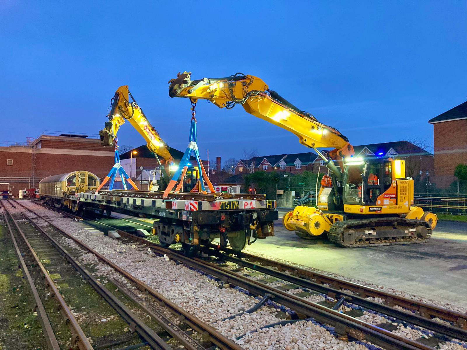 Two yellow rail excavators lifting track equipment beside an engineering train